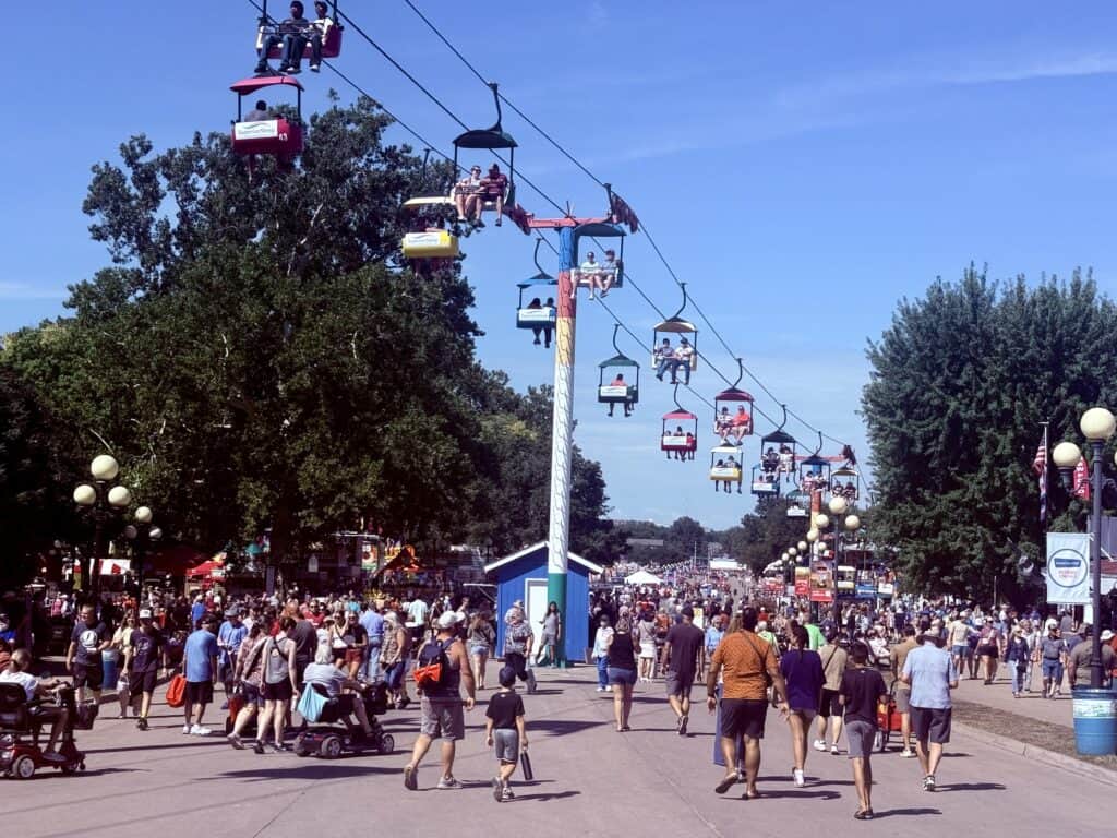 Large crowds of people wander through the fair, with a sky-ride above.