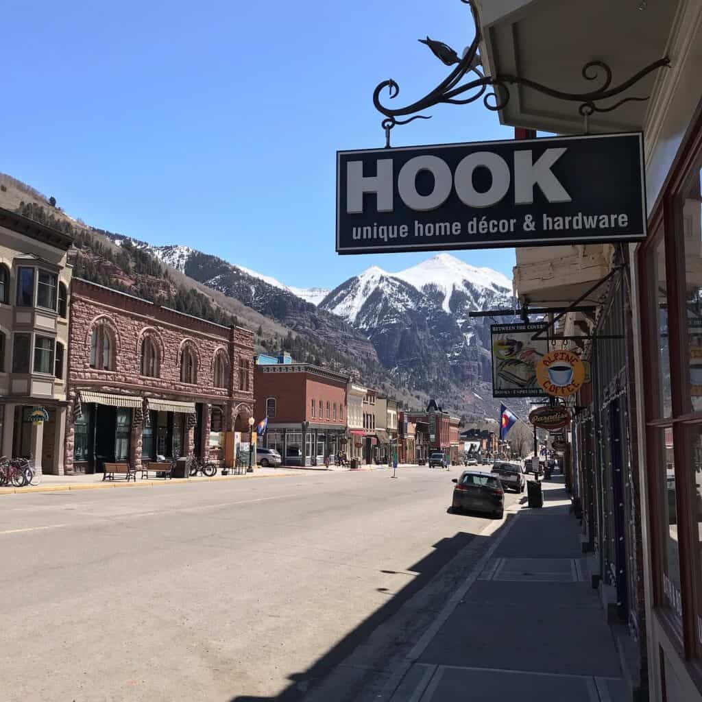 The main shopping street in Telluride, with a sign for Hook, a home decor shop in the foreground.  There are lots of other signs in the background.  At the end of the street are snow-capped mountain peaks.