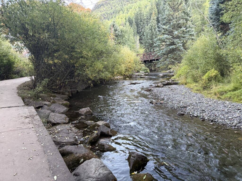 A curving paved trail next to the river lined with big rocks lining one side and small rocks on the other.  There is a bridge crossing the river in the distance, and abundant pine trees and other green trees all over.