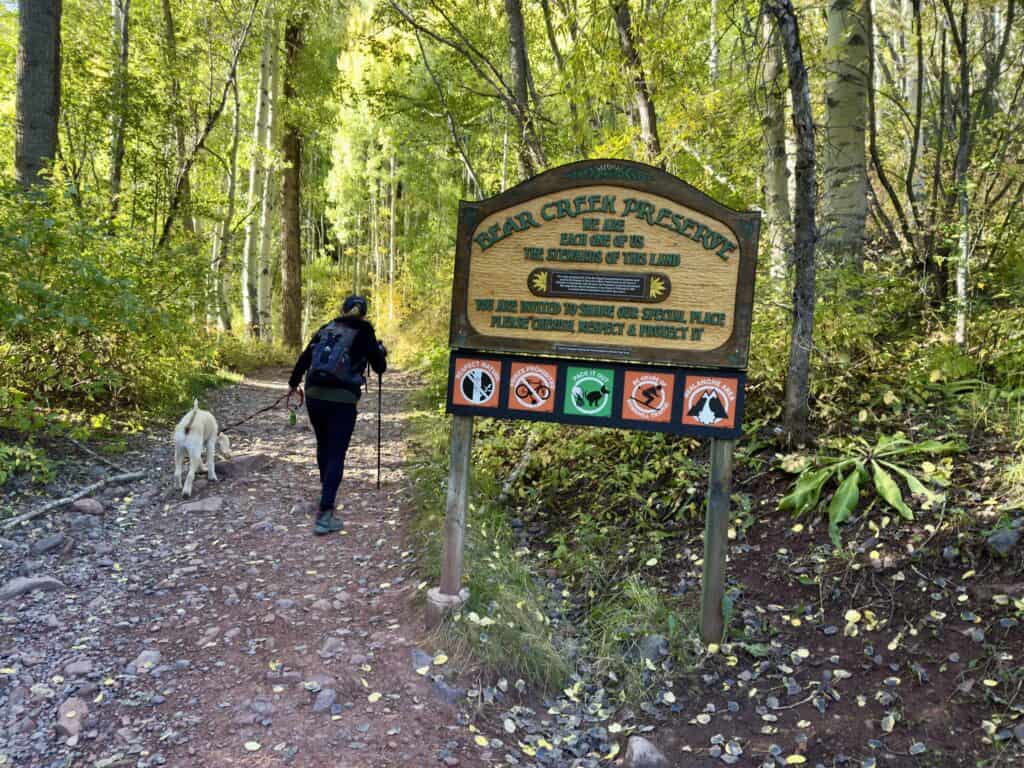 Lisa enters the trail (with trekking poles) next to a big sign announcing the Bear Creek Preserve.  Frisco the Werthwhile Pup, walks beside her.