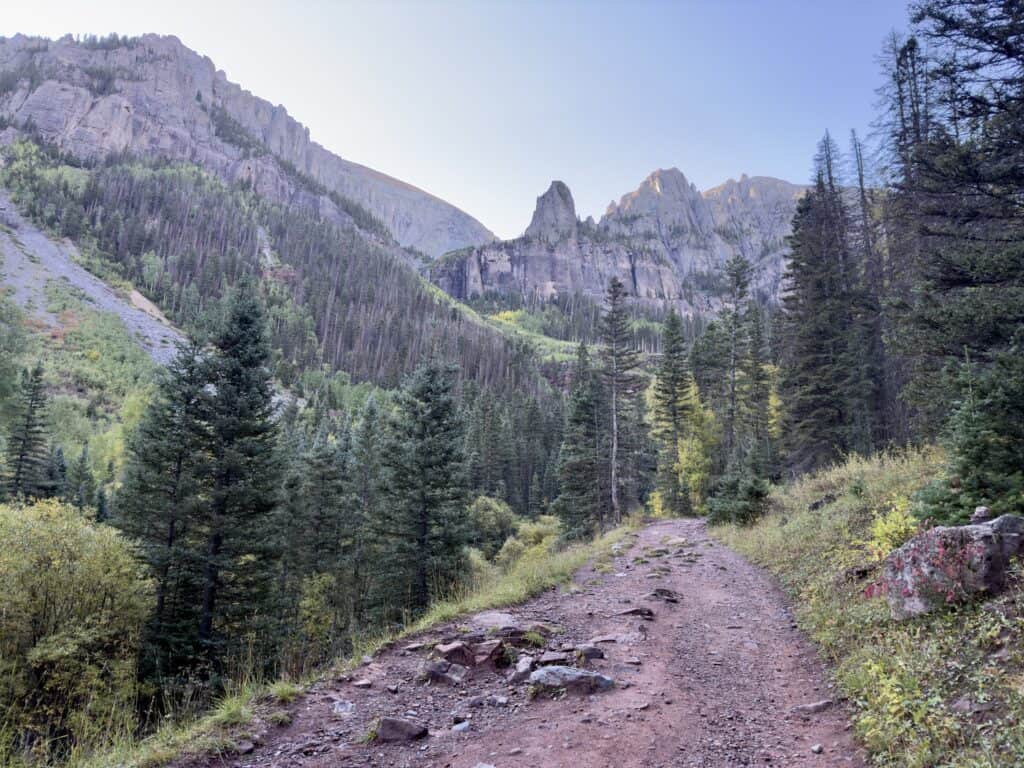 A wide rocky trail with pine trees on both sides and the craggy mountains in the distance.