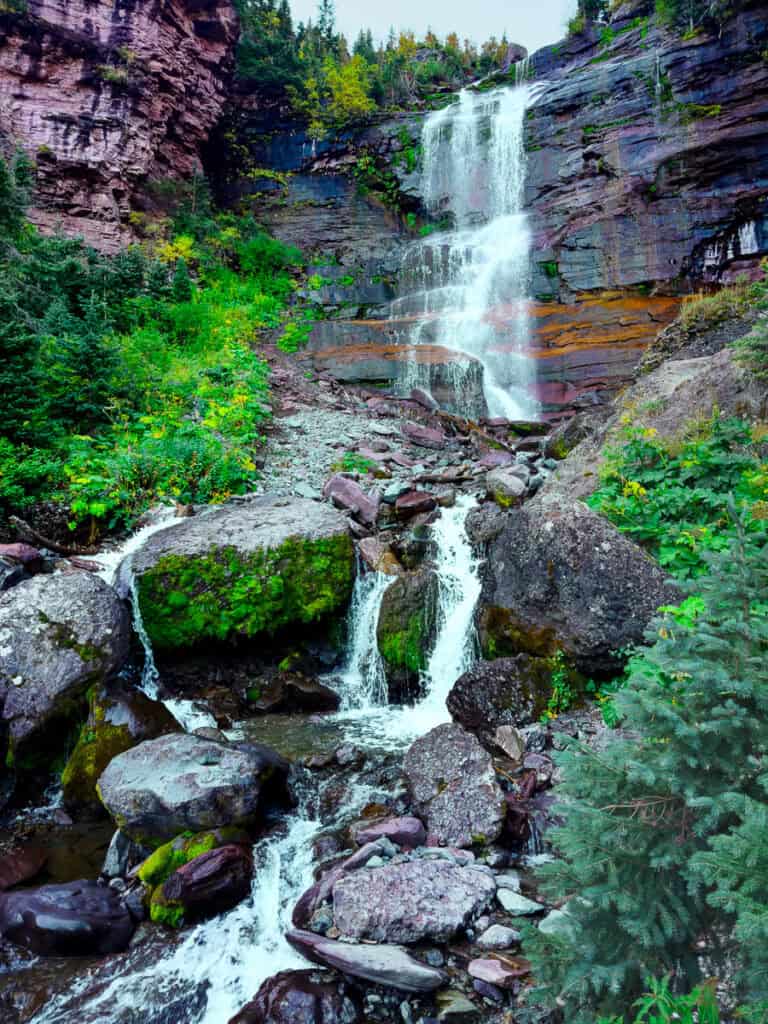 A beautiful, multi-tiered falls flowing down colorful rock, surrounded by green plants and trees.