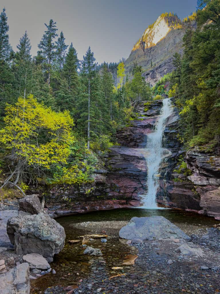 The first falls pour over ledge rock into a crystal clear pool below. Trees line the falls on both sides.