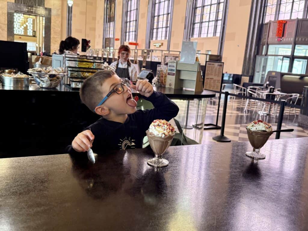 A little boy eating the maricino cherry from atop his chocolate ice cream sundae.