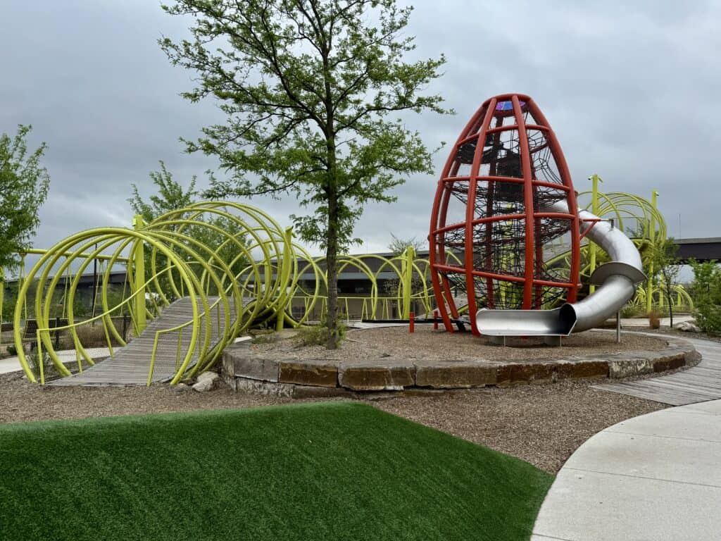 A modern, colorful playhard has unusual shapes to climb in and around and a rocket shaped structure to climb inside and come down a long, twisting slide.