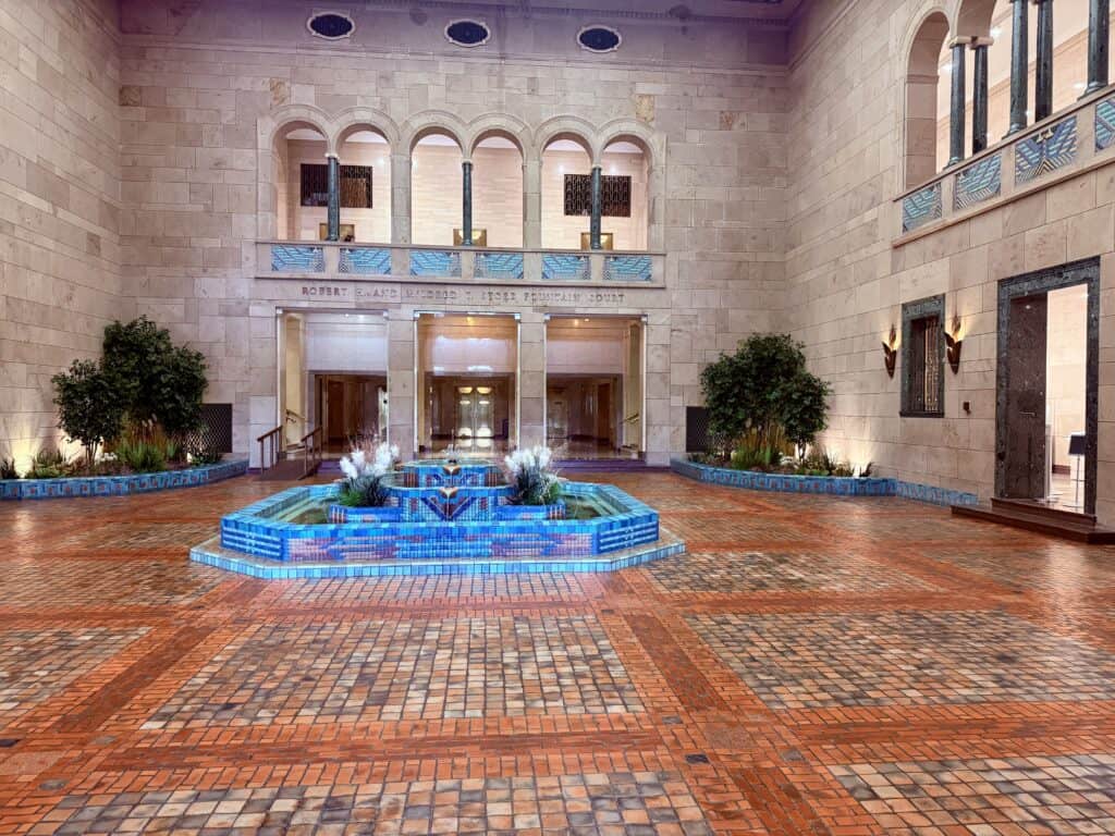 A blue fountain rises from a multi-colored tile floor.  Other than the fountain and some greenery in the corners, the courtyard is open space.  Lines of arches on the upper floor look down over the courtyard.