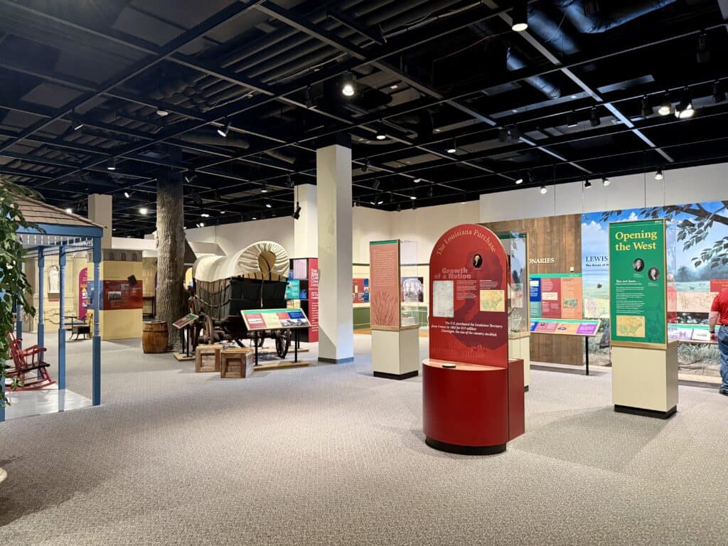 A room full of exhibits, including a full-sized covered wagon, information about Lewis and Clark, and old rocking chairs sitting on the porch of a storefront.