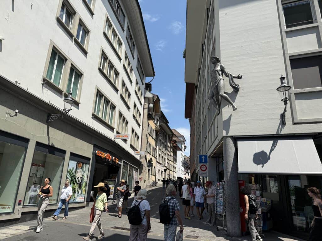 A dozen or so visitors wander tha narrow cobblestone street lined with multi-story buildings.  A bronze statue of a woman hangs off a building at about the third story.