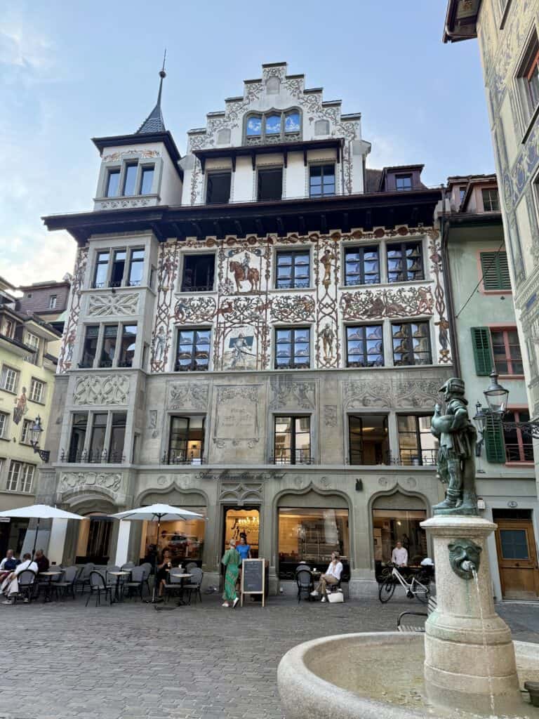 Midieval 5-story buildding with frescos on the outside.  It is located in a plaza with a restaurant with outside seating and a fountain.