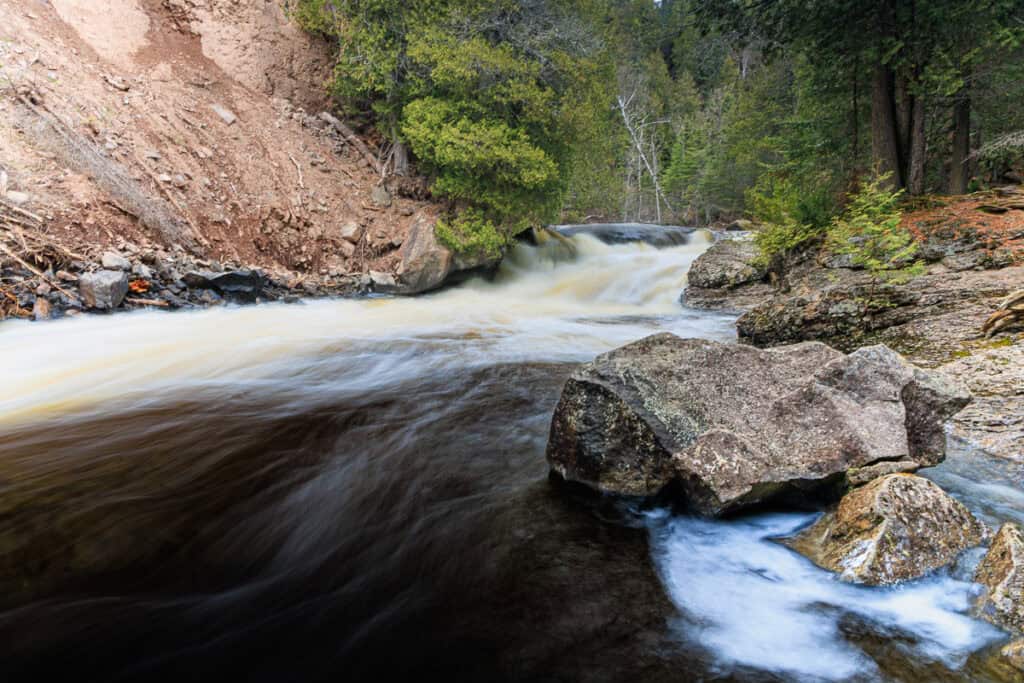 Small but beautiful waterfall in the background with the river flowing by.  Tall rocky walls and big boulders surround the river.