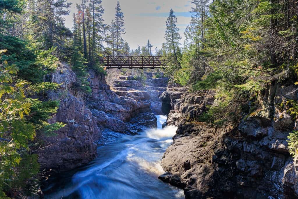 Idyllic scene with a pretty wooden bridge arched over the deep cavern and one of the falls roaring down beneath it.