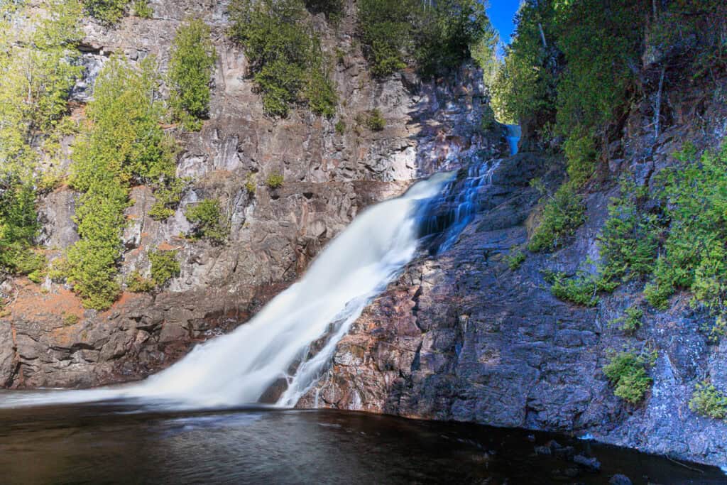 A silky waterfall appears from between two steep rock walls and pours down to the river below.