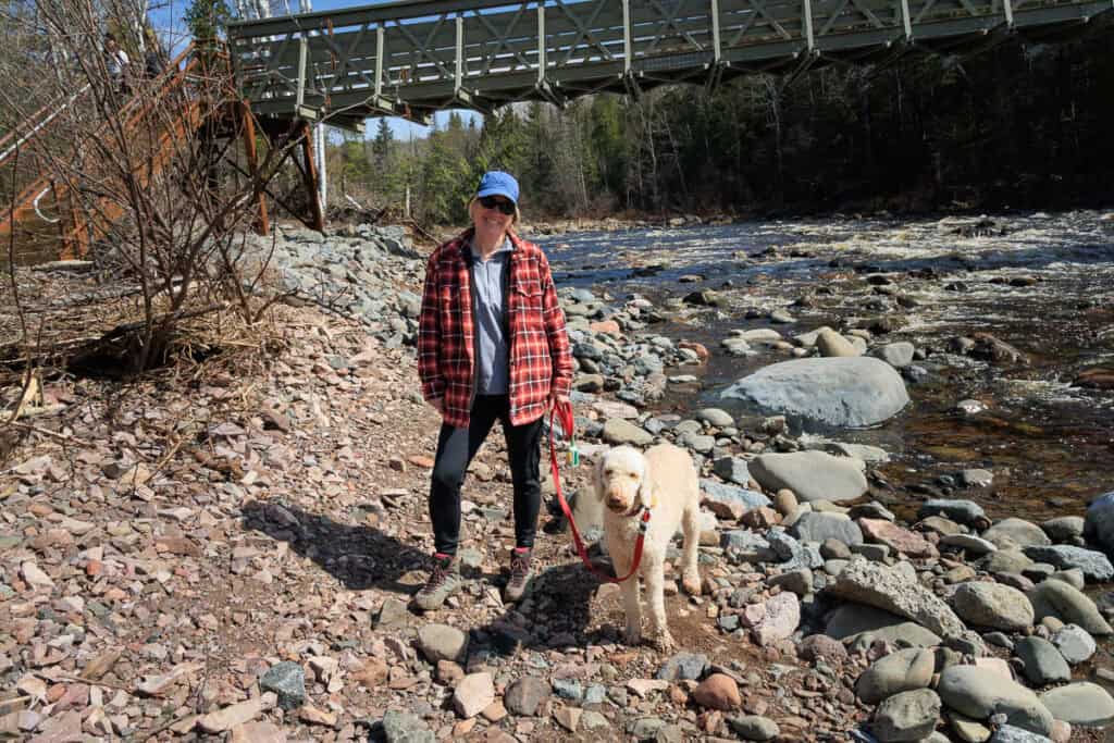 A smiling Lisa and her dog stand beneath the pedestrian bridge over the river, which is flowing quickly and full of rocks and boulders.