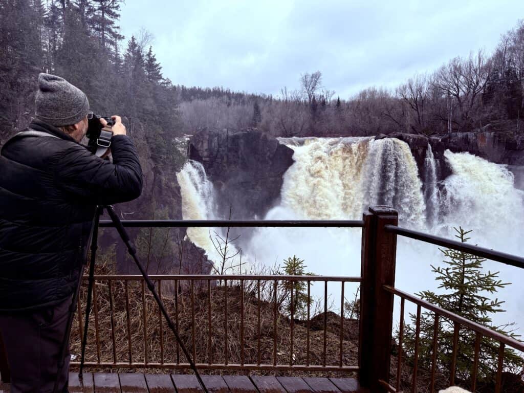 Jeff is taking photo of the falls  from one of the overlooks with rails on all sides.  He has a tripod and his camera, and is dressed in a coat and hat.