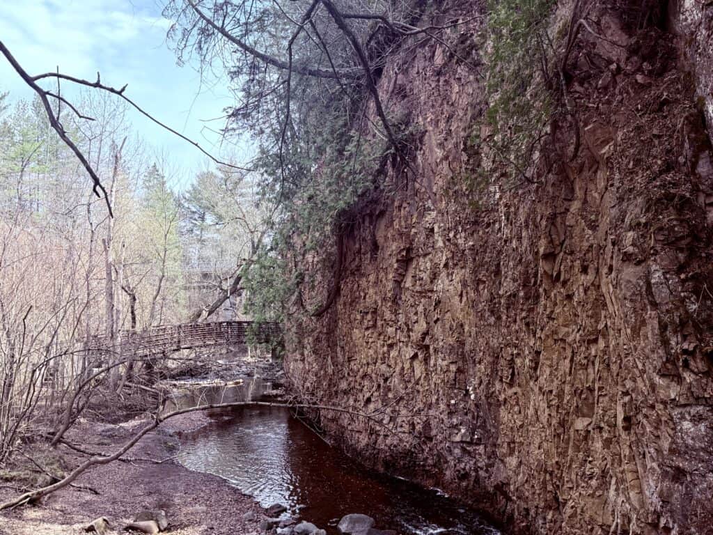A bridge passes over a stream next to a tall red-rock cliff.