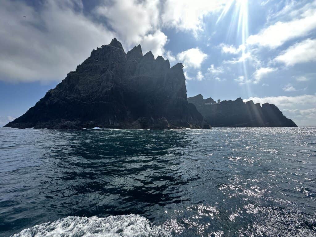 Craggy rocks in the sea, making up the Skellig Michael.  The sun shines through the puffy white clouds above.